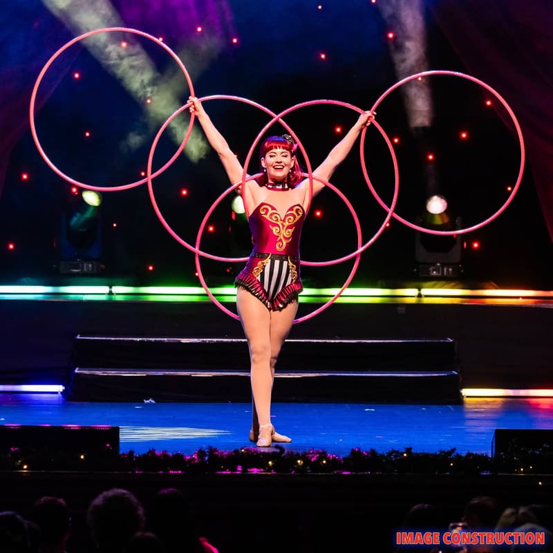 Performer in red sequined costume manipulating illuminated hoola hoops on stage under purple and blue lighting.