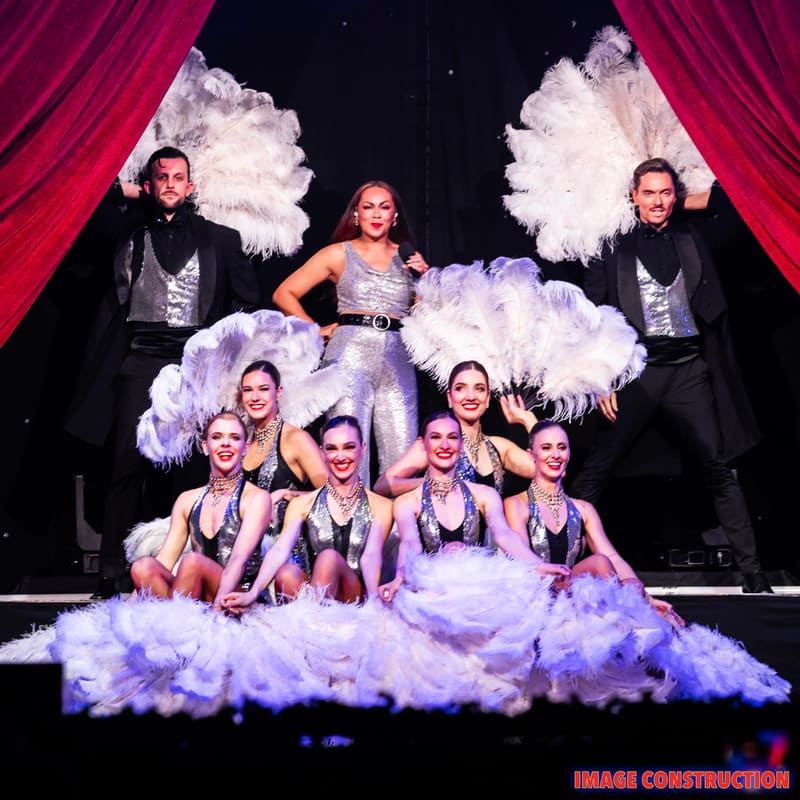 Performers in elaborate costumes with oversized white feather headdresses pose on stage beneath purple-lit curtains during a theatrical production.