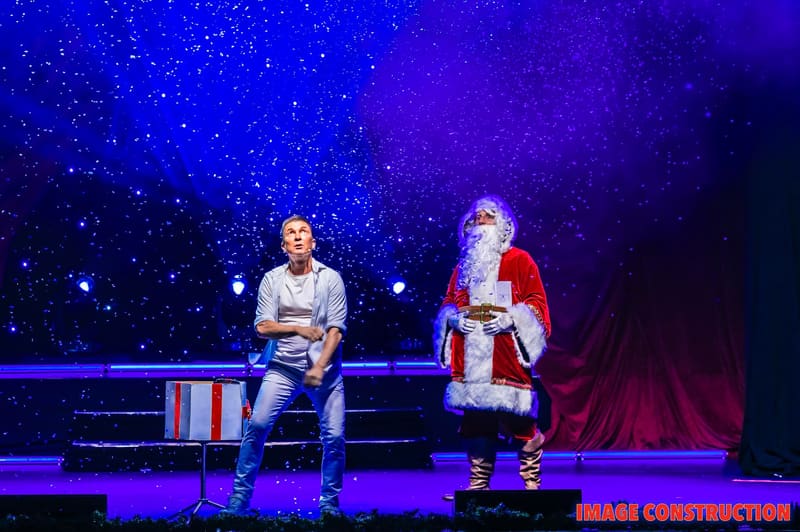 A person in casual clothing sits beside Santa on stage against a starlit backdrop at The Christmas Spectacular.