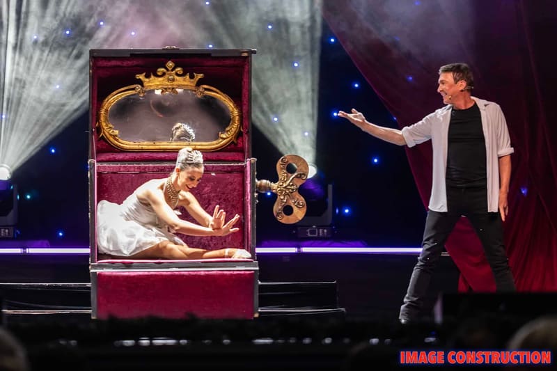 Performer gestures toward an ornate display case containing a reclining figure in white, lit by theatrical spotlights.