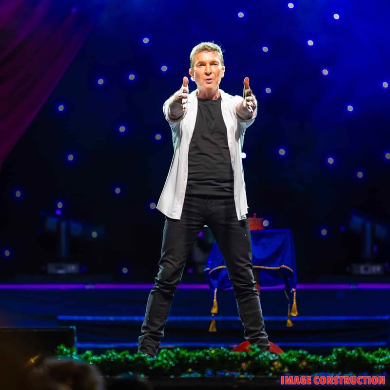 Performer in white blazer and black shirt gestures enthusiastically on stage amid blue spotlights and festive greenery.