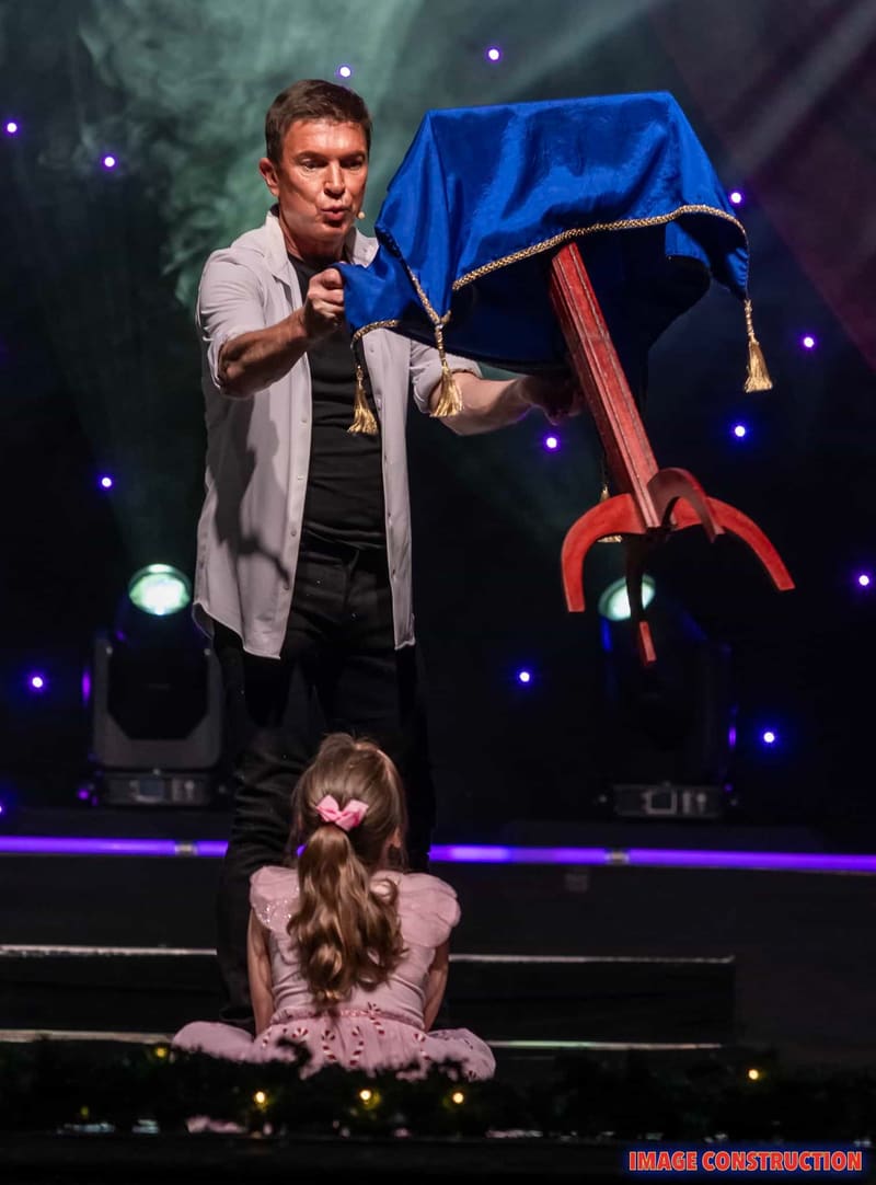 Performer holds microphone beside large red mechanical claw under blue fabric during Christmas Spectacular stage show.