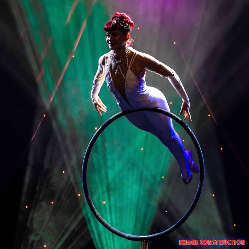 Performer balanced on a large hoop mid-air against vibrant green and purple stage lighting.