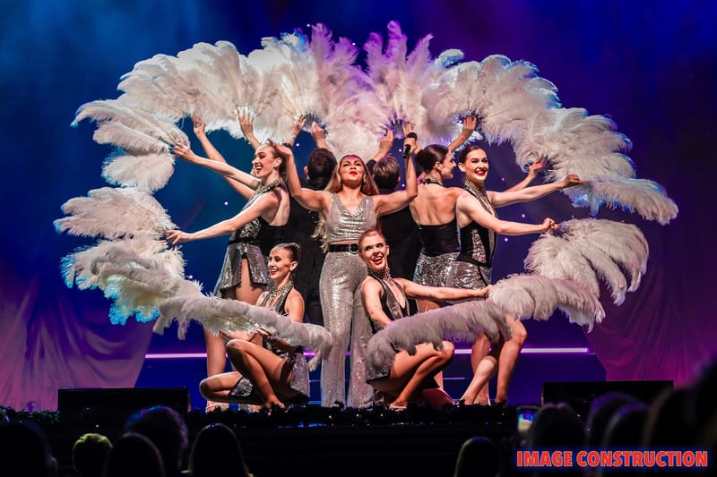 Ensemble of performers in sequined costumes posed with large white feathered wings under purple stage lighting during The Christmas Spectacular.