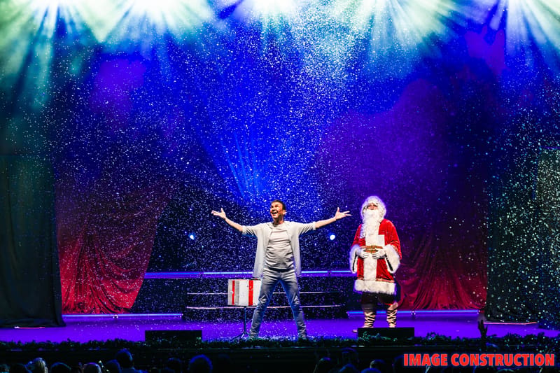 Two performers on stage with arms outstretched amid falling snow and vibrant blue and purple lighting during The Christmas Spectacular.