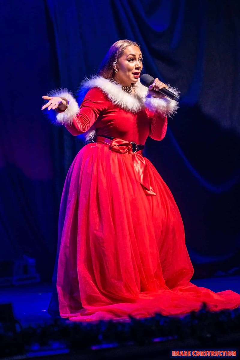 Performer in a red velvet gown with white fur trim sings into a microphone under blue stage lighting.