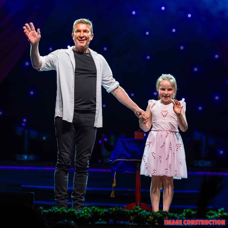 Adult and young child hold hands on stage, waving to audience under blue starlit backdrop.