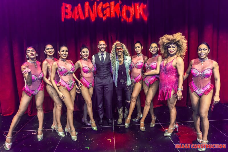 Cast of The Ladyboys of Bangkok pose together on stage in sequined costumes beneath a neon Bangkok sign.