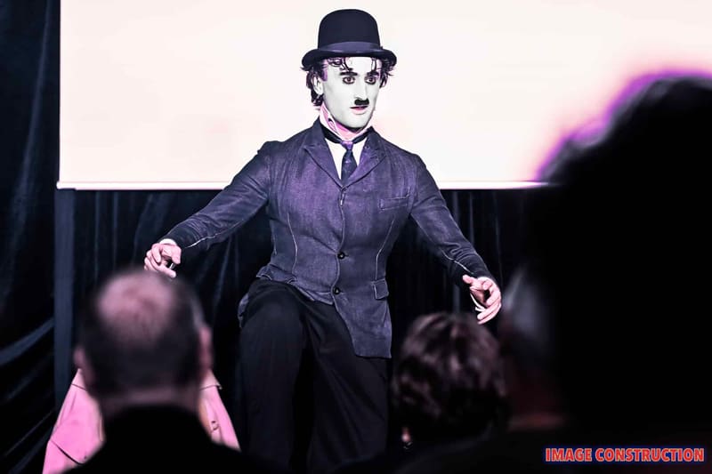 Performer in Chaplin's iconic bowler hat and suit gestures expressively on stage, bathed in purple spotlight lighting.