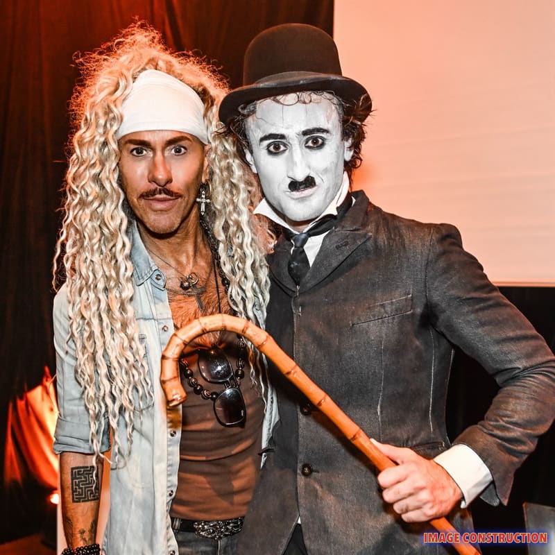 Performer in curled blonde wig and headband poses beside Charlie Chaplin figure in bowler hat and suit, holding a cane.
