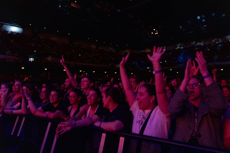 Crowd of audience members with hands raised, illuminated by purple and pink stage lighting at a live concert.