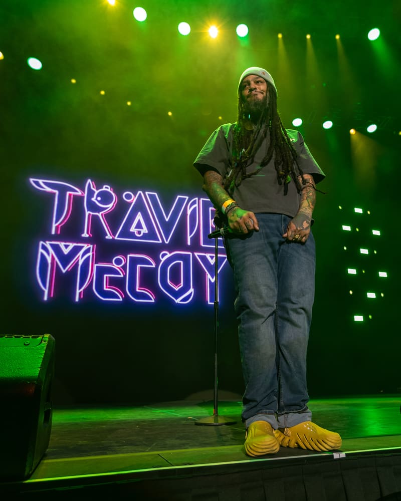 Performer on stage under green and white stage lights, standing beside a neon "TRAVIE McCOY" sign.