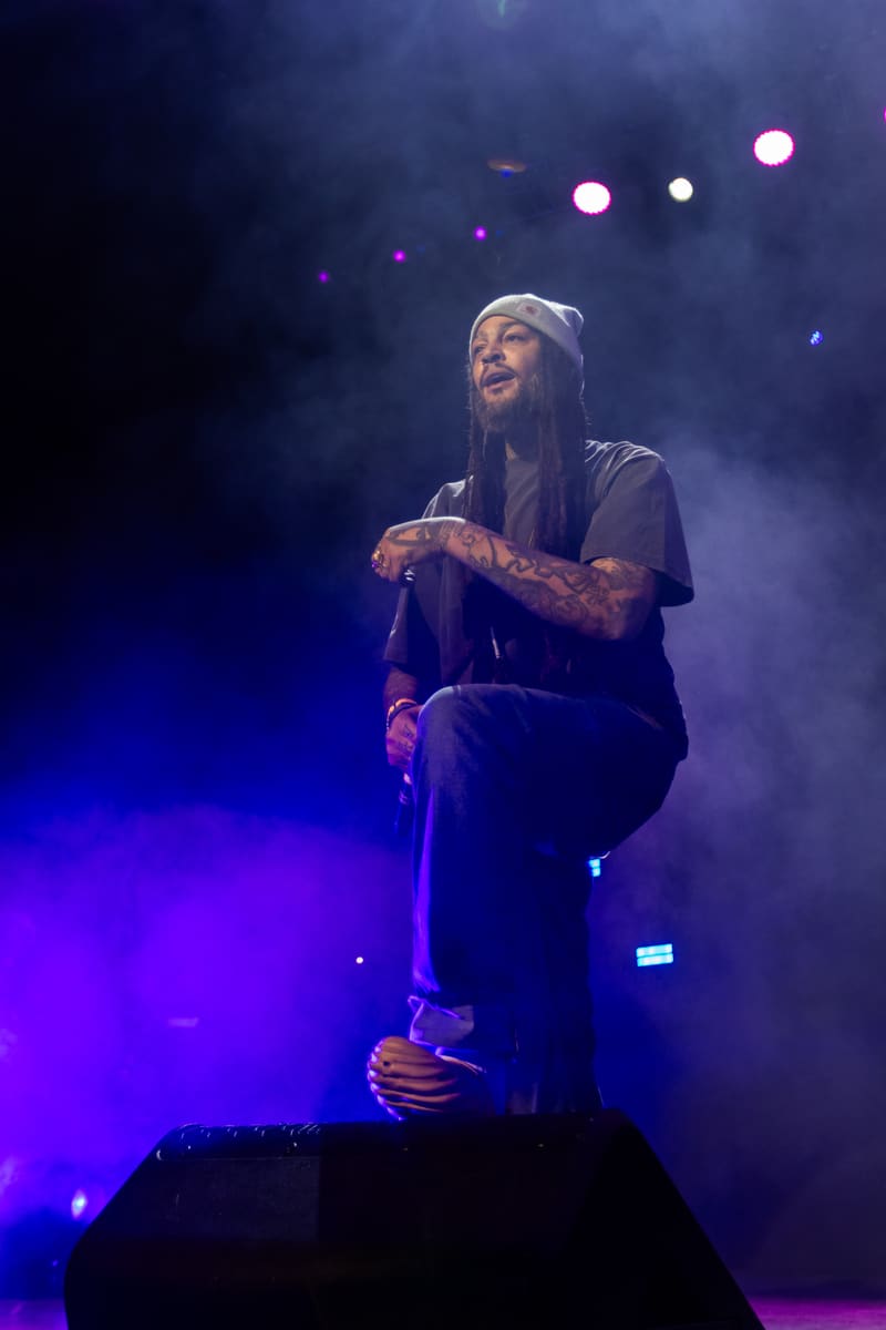 Performer on stage holding microphone, bathed in blue and purple stage lighting with atmospheric smoke and spotlights.