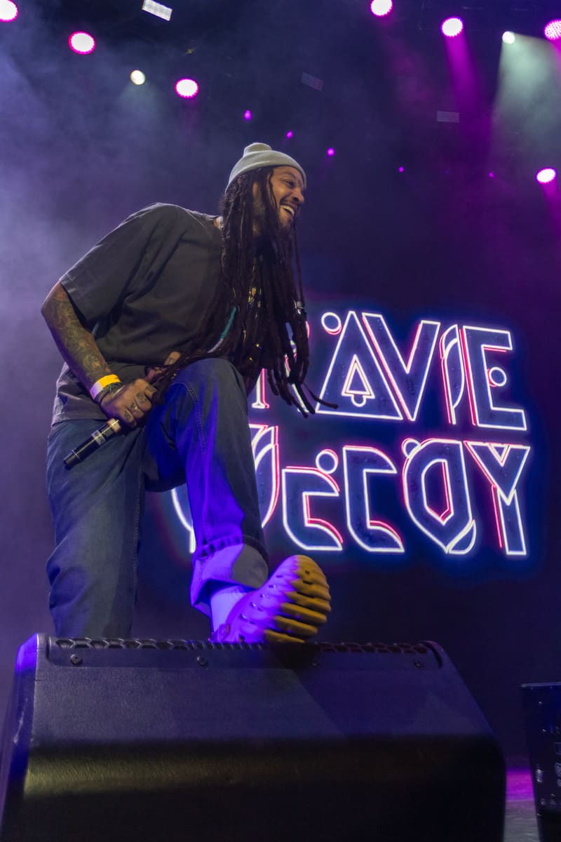 Performer in a white hat and black shirt stands on stage under purple lighting during Fridayz Live.