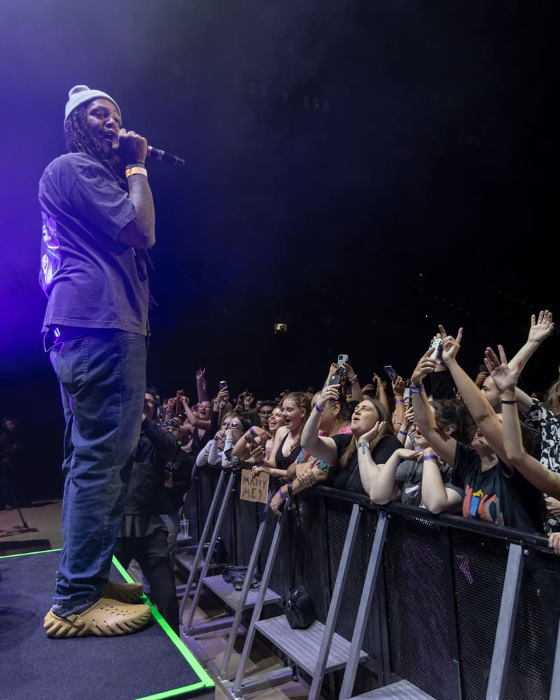 Performer on stage with microphone, bathed in purple light, facing an engaged crowd with raised hands at a live music event.