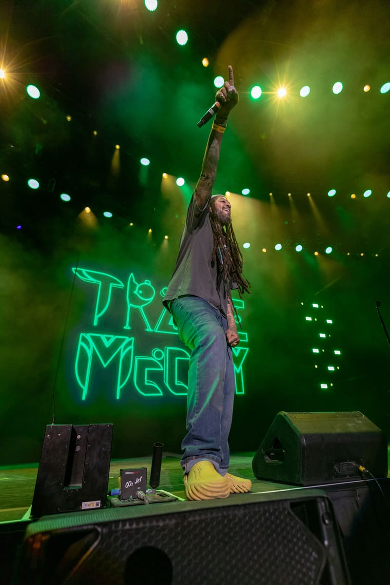 Performer stands on stage with arm raised, singing into microphone, with neon green branding and stage lights behind.