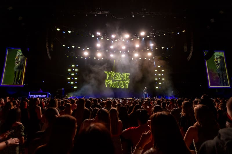 Crowd of concertgoers with hands raised facing a brightly lit stage displaying "TRAVIE McCOY" text.