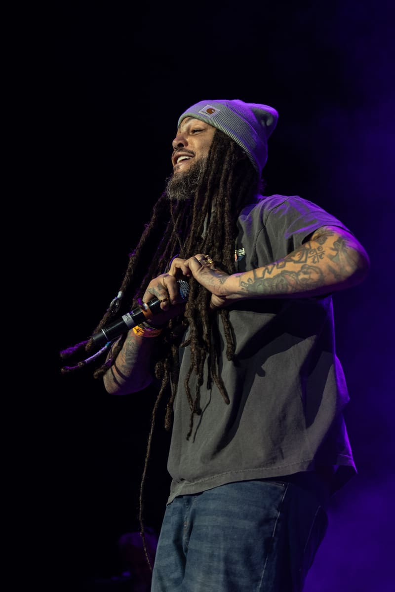 Performer with dreadlocks and beanie holding microphone on stage under purple lighting during Fridayz Live.