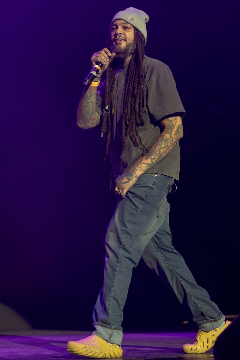 Performer on stage holding a microphone, wearing a beanie and dreadlocks, illuminated by purple stage lighting.
