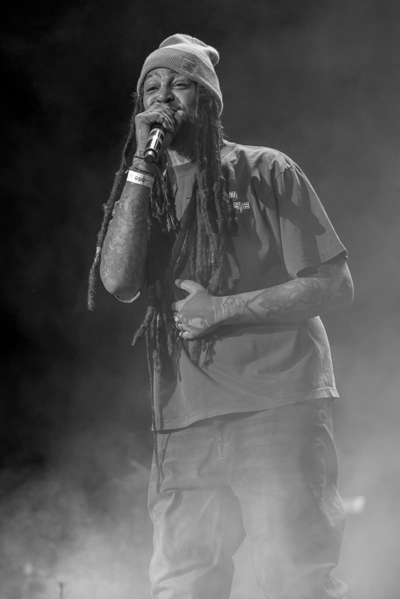 Performer singing into microphone on stage, wearing a beanie and casual shirt, surrounded by atmospheric smoke.