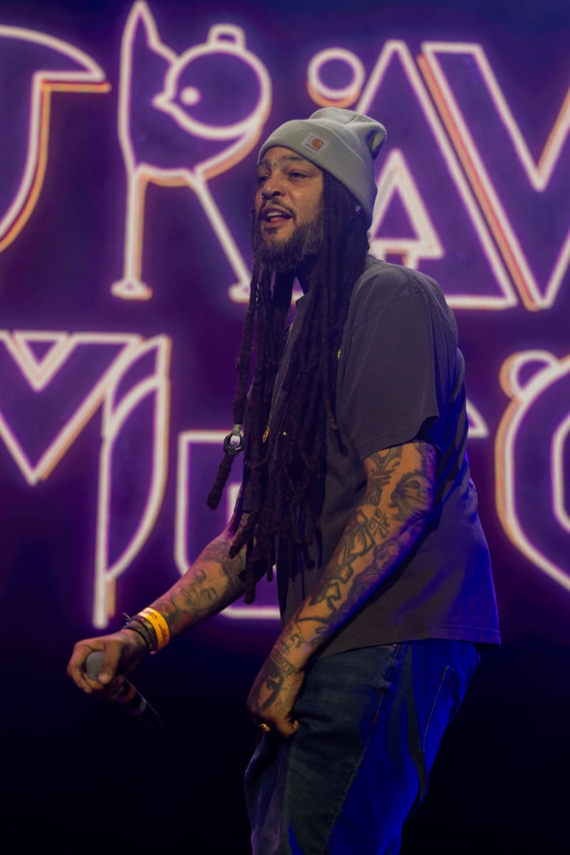Performer with dreadlocks and grey beanie wearing tattooed arms on stage against purple neon signage.