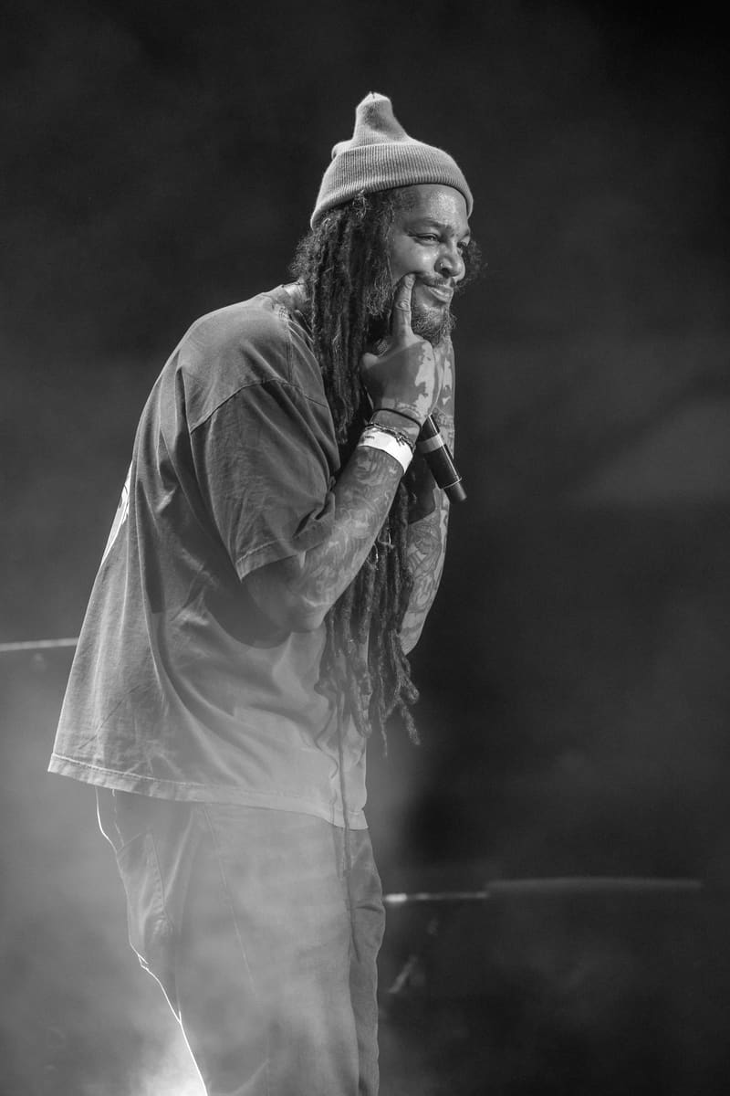 Performer with long dreadlocks and beanie singing into microphone on stage in black and white.