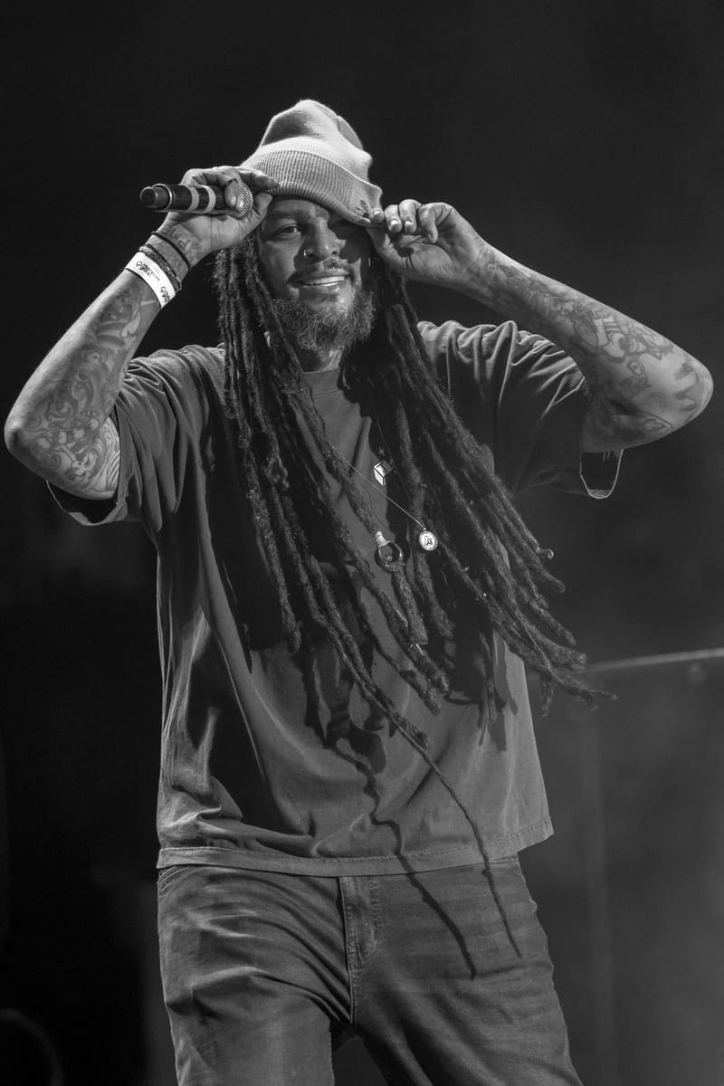 Performer on stage adjusting a white fedora hat and holding a microphone, photographed in black and white against a dark background.