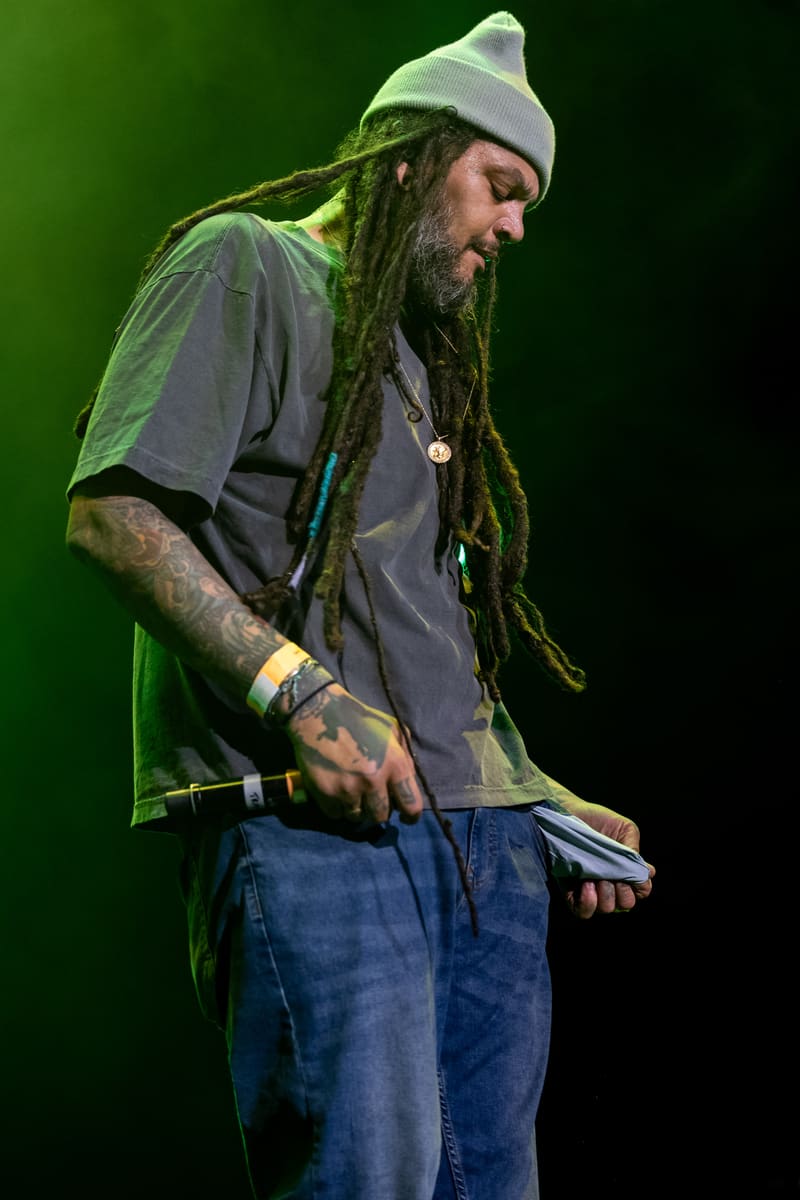 Performer with dreadlocks and beanie hat playing guitar on stage under green lighting.