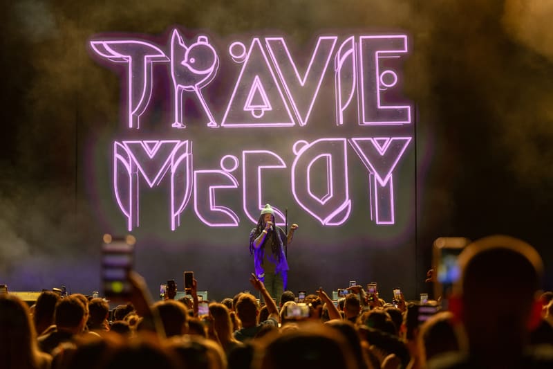 Performer on stage beneath purple neon "TRAVIE McCOY" signage, surrounded by audience members with raised hands.