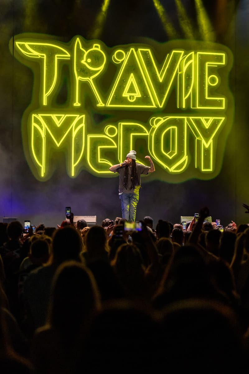 Performer on stage beneath a massive yellow neon "TRAVIE McCOY" sign, surrounded by a crowd of audience members.
