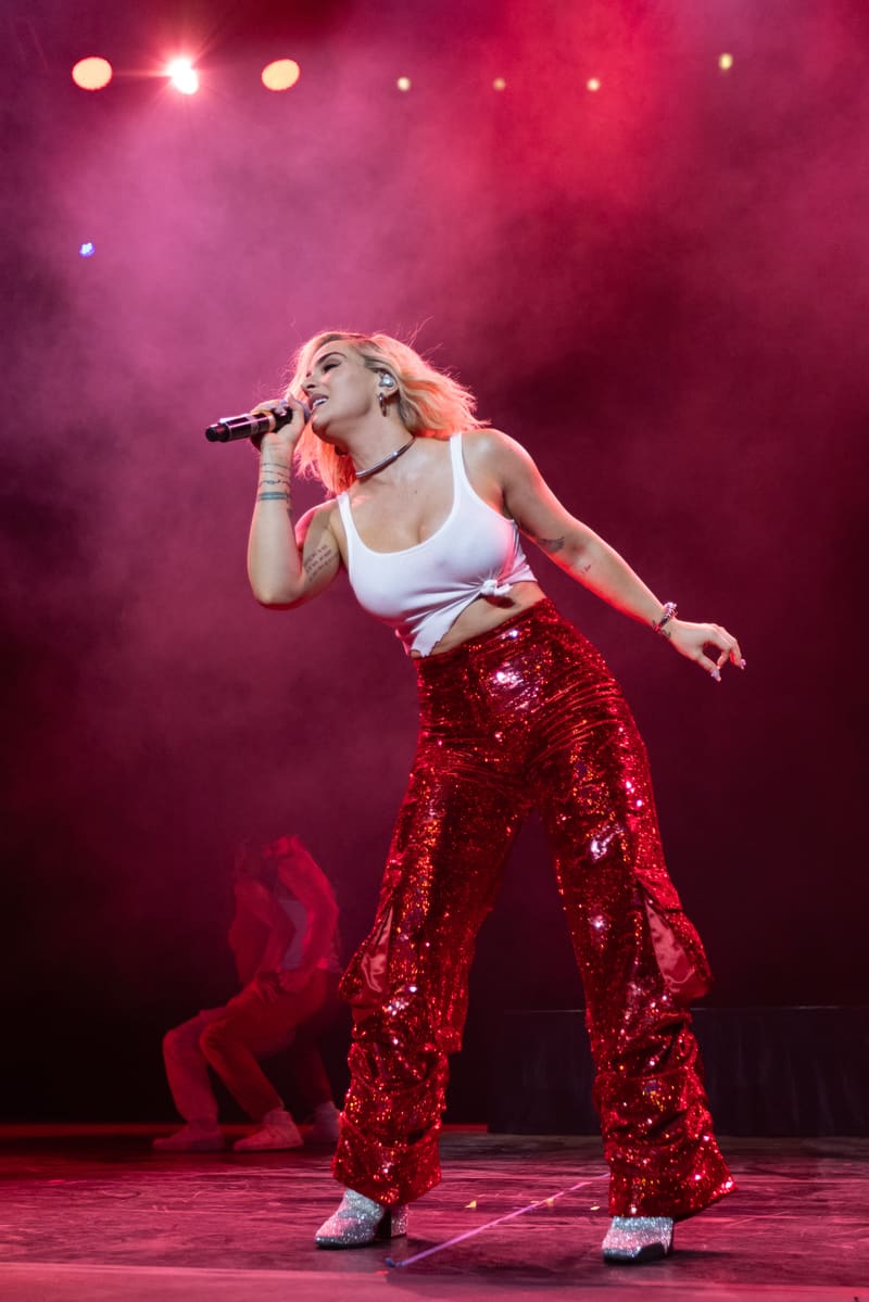 Performer singing into microphone wearing white crop top and sequinned red pants, bathed in pink stage lighting.