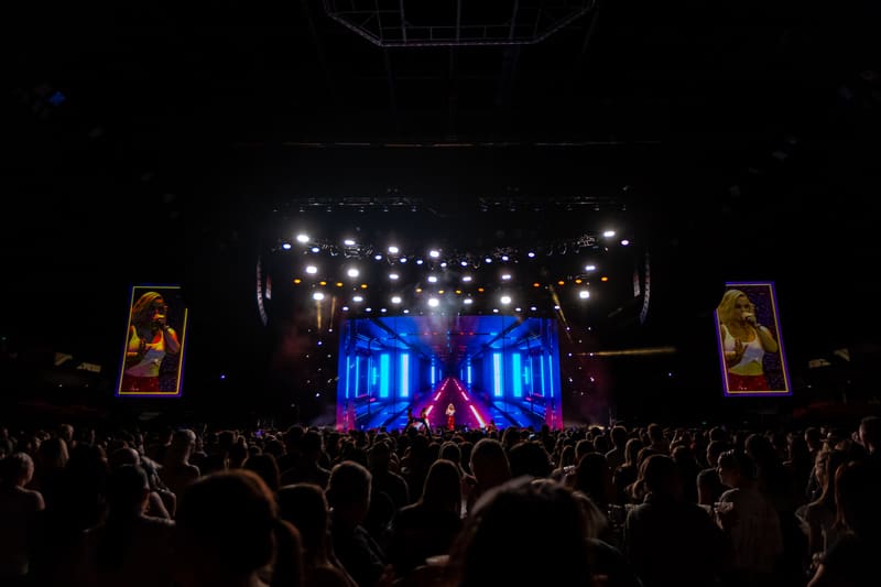 Crowd of concertgoers with hands raised facing illuminated blue stage structure under bright overhead lights.