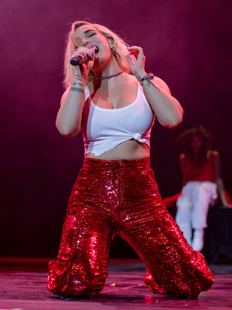 Performer singing into microphone on stage in white crop top and red sequined trousers, bathed in pink stage lighting.