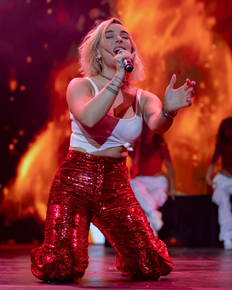 Performer singing into microphone wearing red sequined pants and white crop top against warm-toned stage lighting.