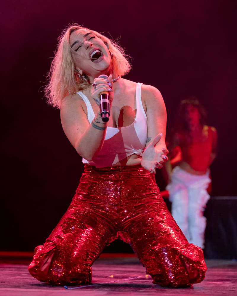Performer singing with microphone on stage, wearing red sequinned pants and white top, bathed in pink and red stage lighting.