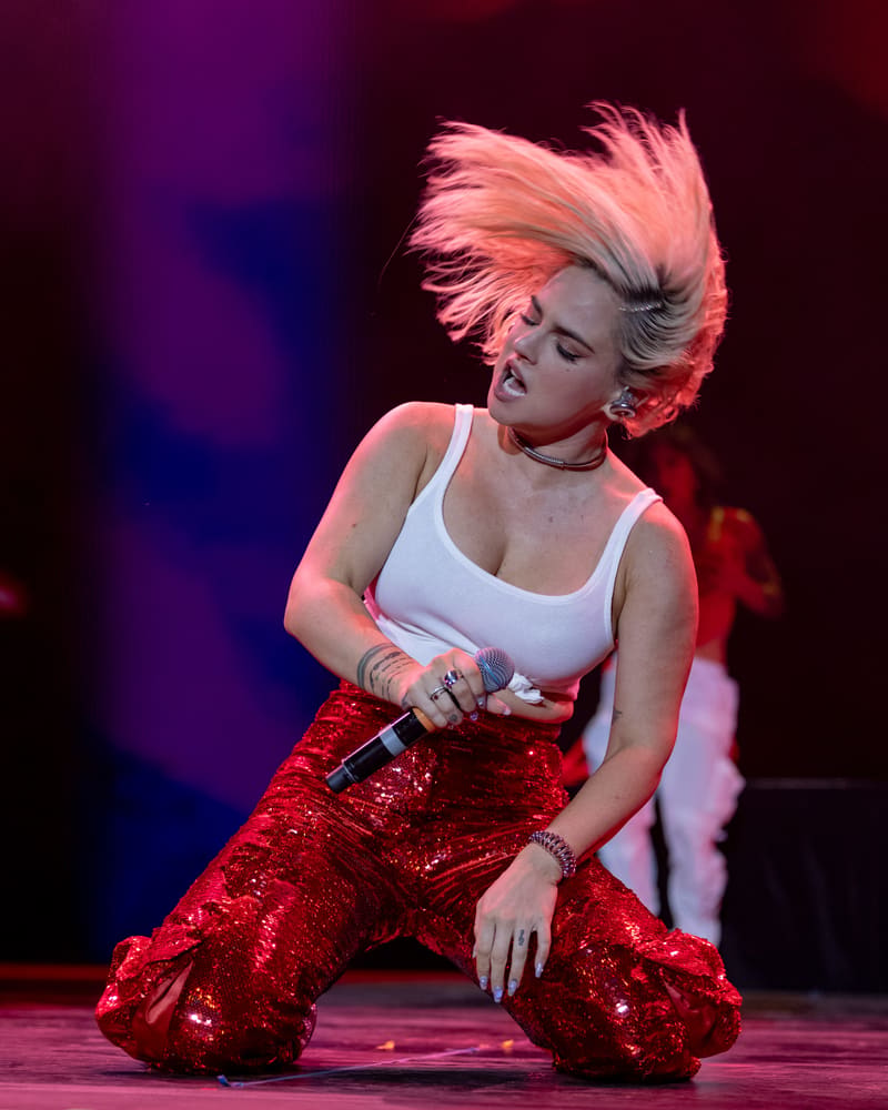 Performer in white tank top and red sequinned pants kneels on stage, illuminated by purple and red spotlight lighting.
