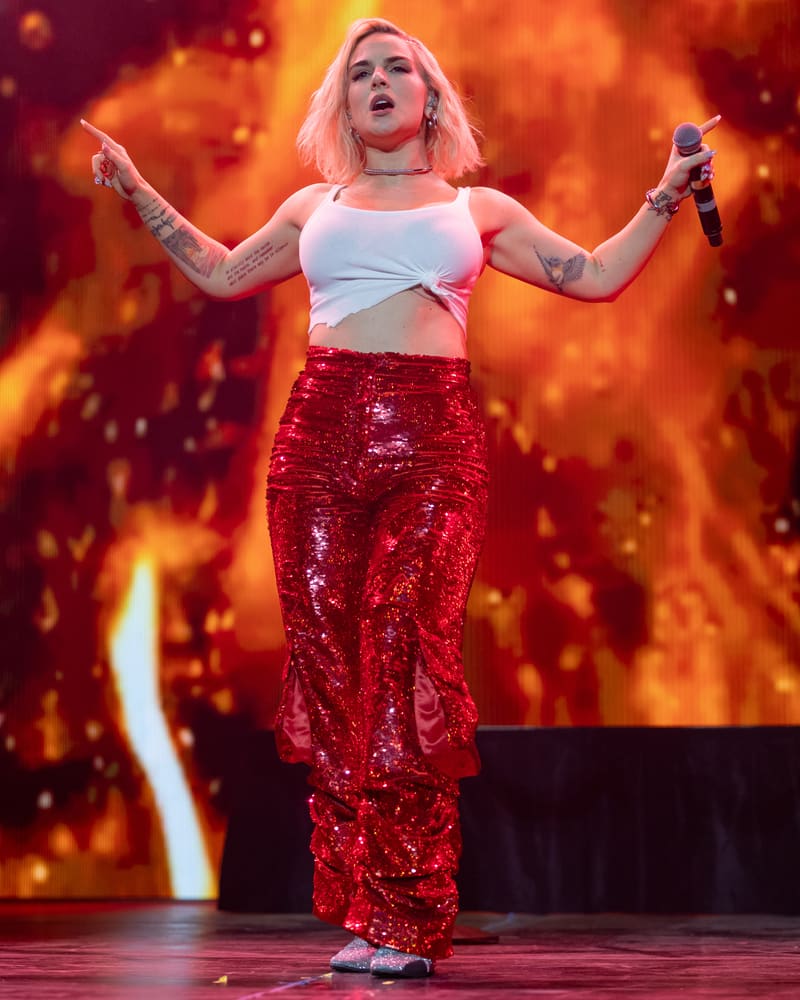 Performer in white crop top and red sequined trousers holds microphone on stage against fiery orange backdrop.