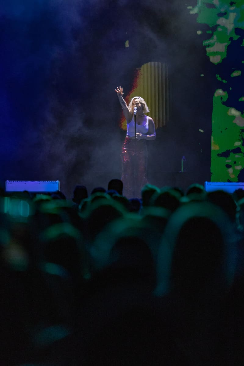 Performer on stage bathed in blue and green stage lighting, singing to an engaged audience in darkness.