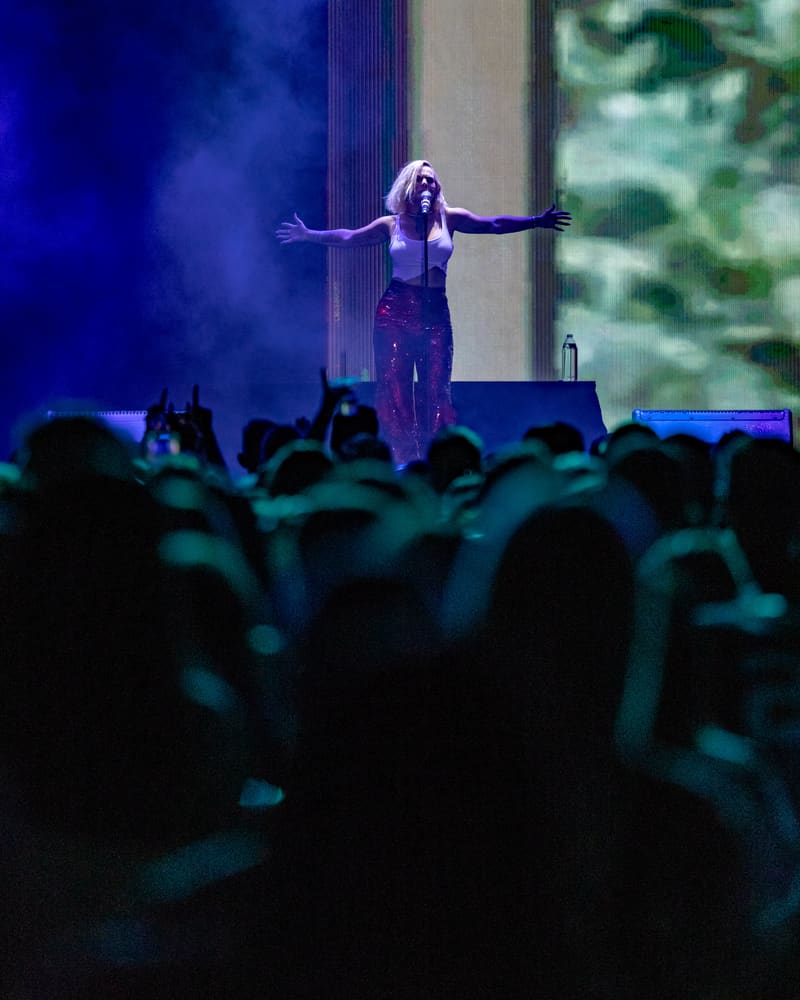 Performer on stage with arms outstretched, lit by blue spotlights, facing an engaged audience.