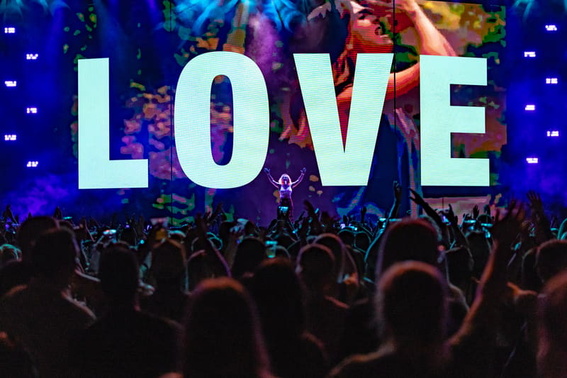 Large "LOVE" letters illuminated on stage above a crowd of concertgoers with hands raised during a live music performance.