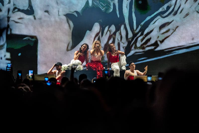 Group of performers on stage under warm spotlights with large-scale projected imagery behind them during Jojo – Fridayz Live.