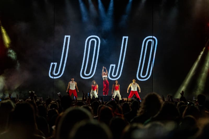 Four performers in red outfits on stage beneath glowing "JOJO" neon sign, audience in foreground.