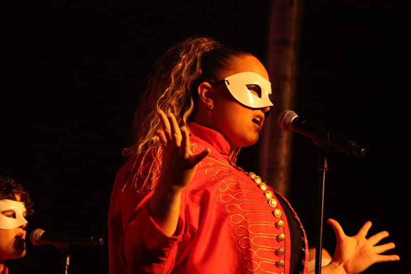 Performer in a gold mask and red sequinned jacket gestures expressively at a microphone under warm stage lighting.