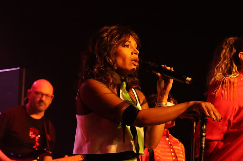 Performer in colourful beaded jewellery gestures expressively at microphone under warm stage lighting, with band members beside her.
