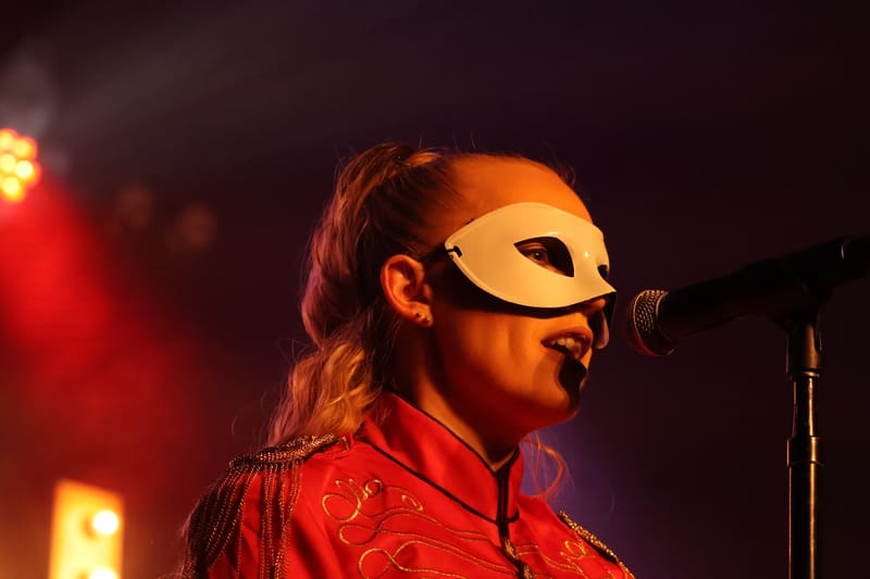 Performer wearing a white mask and red jacket sings into a microphone under warm stage lighting.