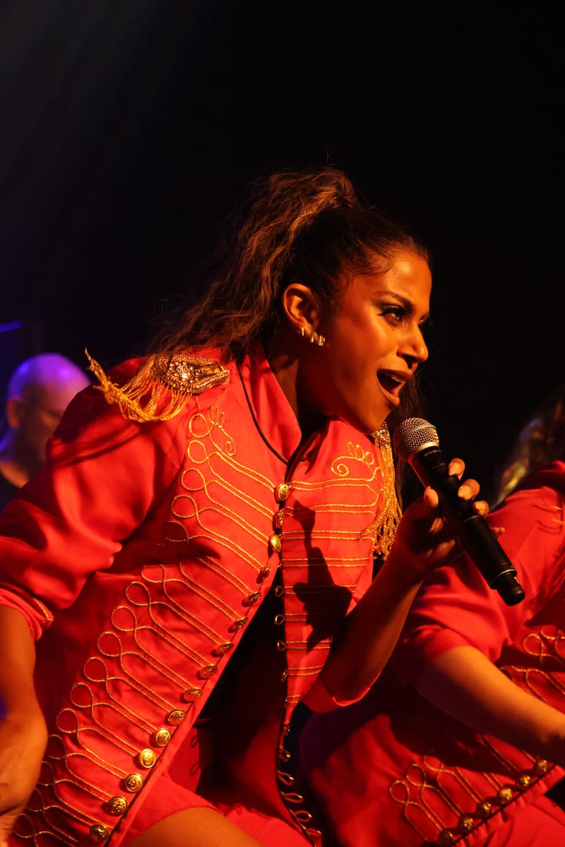 Performer in red sequined jacket and gold jewellery singing into microphone under warm stage lighting.