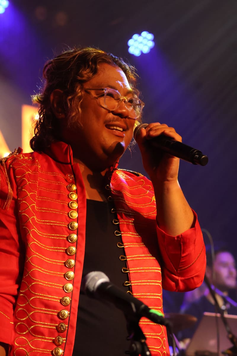 Performer in red military-style jacket with gold buttons singing into microphone under blue and amber stage lighting.
