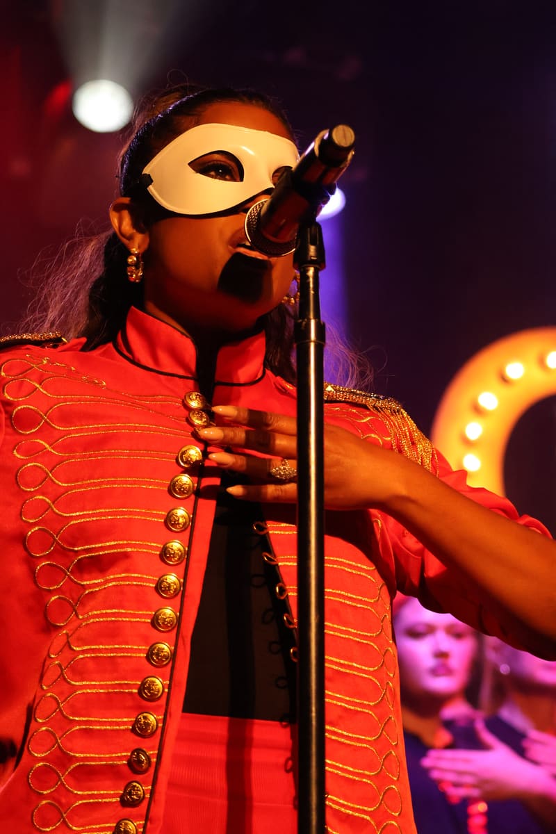 Performer in a gold mask and red sequined jacket sings at a microphone under stage lighting.