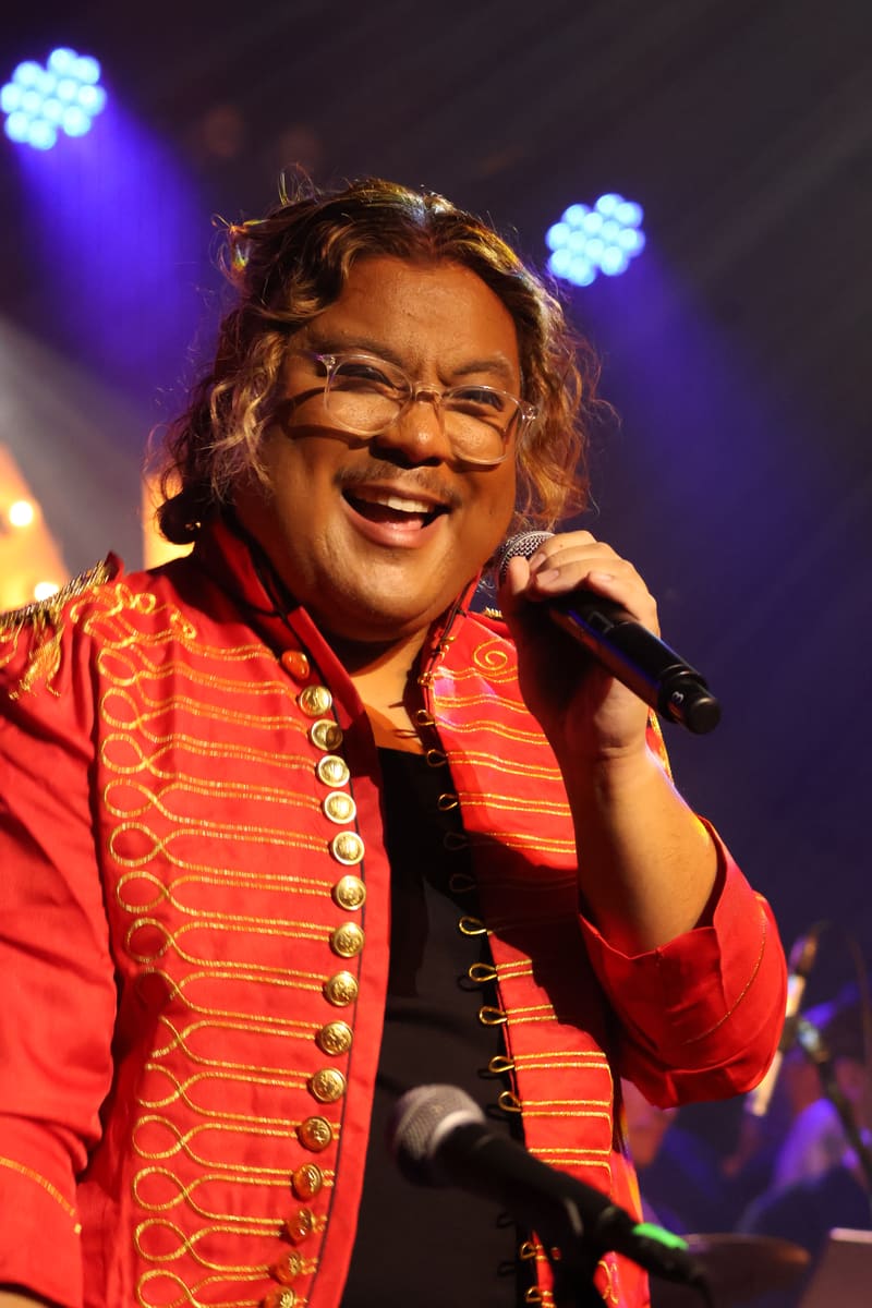 Performer in red embellished jacket singing into microphone under purple stage lighting.