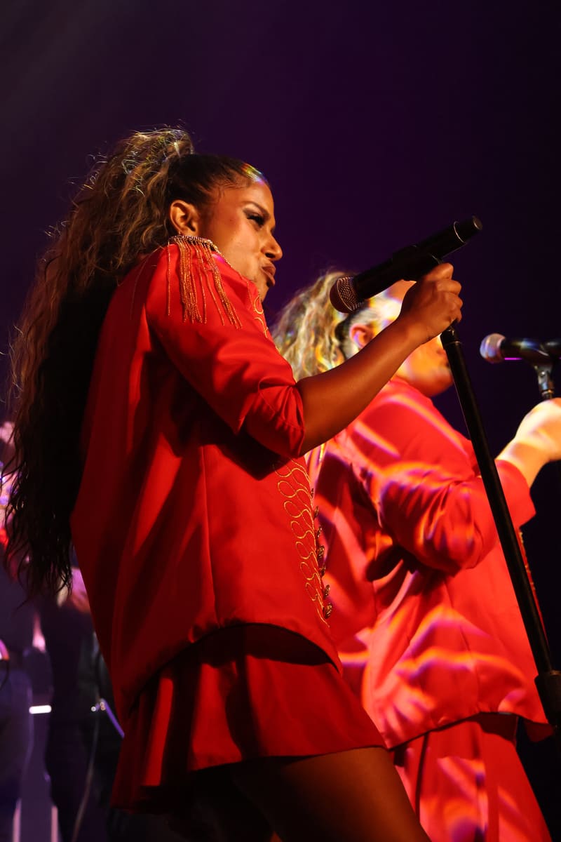 Performer in a vibrant red outfit singing into a microphone on a darkly lit stage.
